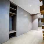 A modern, spacious walk-in closet with built-in grey cabinets and shelves made of luxury bespoke furniture under bright recessed lighting. A wooden staircase with brown steps and a black railing is visible on the right. The floor is made of light-colored wood.