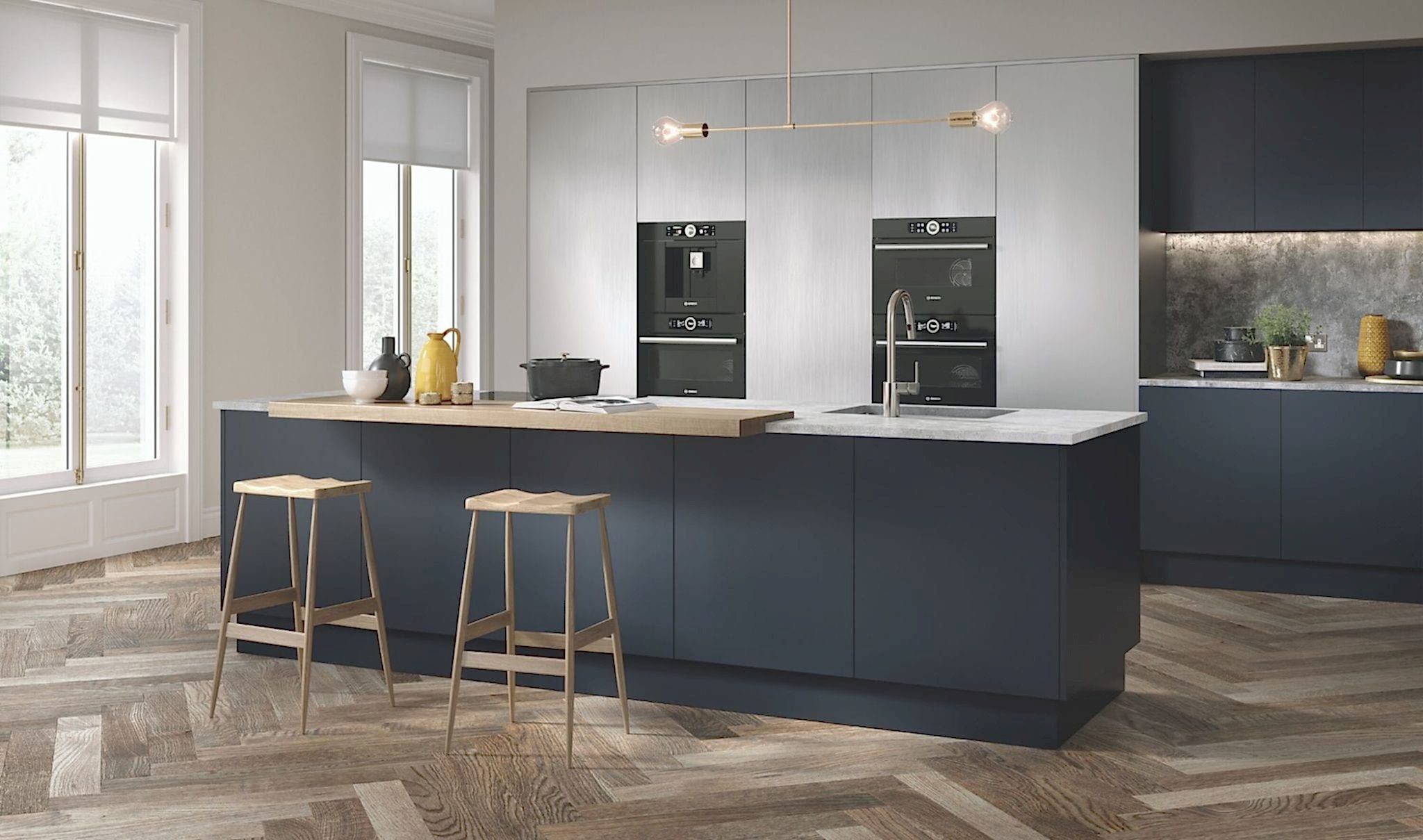 A modern kitchen featuring a large island with a white countertop and two wooden stools. The room has dark blue lower cabinets and light grey upper cabinets. Built-in ovens and a sleek stovetop are visible. Herringbone wood floors add warmth to the space, while a window provides natural light, perfect for contemporary kitchens.