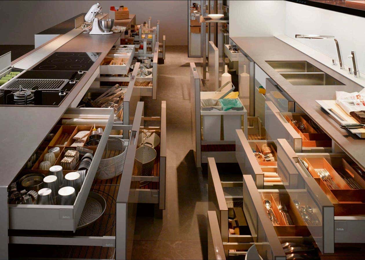 A modern kitchen boasts numerous open drawers showcasing organized utensils, kitchenware, and storage items. The space features a sleek countertop with a stove, a double sink, and a mixer on the counter. Designed for optimal storage efficiency, this kitchen epitomizes contemporary kitchens.