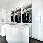 A modern walk-in closet featuring glossy white cabinetry, open shelves, and abundant hanging space for clothes. Two white islands are centered in the room, offering extra storage and surface area. This elegant space integrates seamlessly with bedroom furniture and boasts wooden flooring and ample lighting.