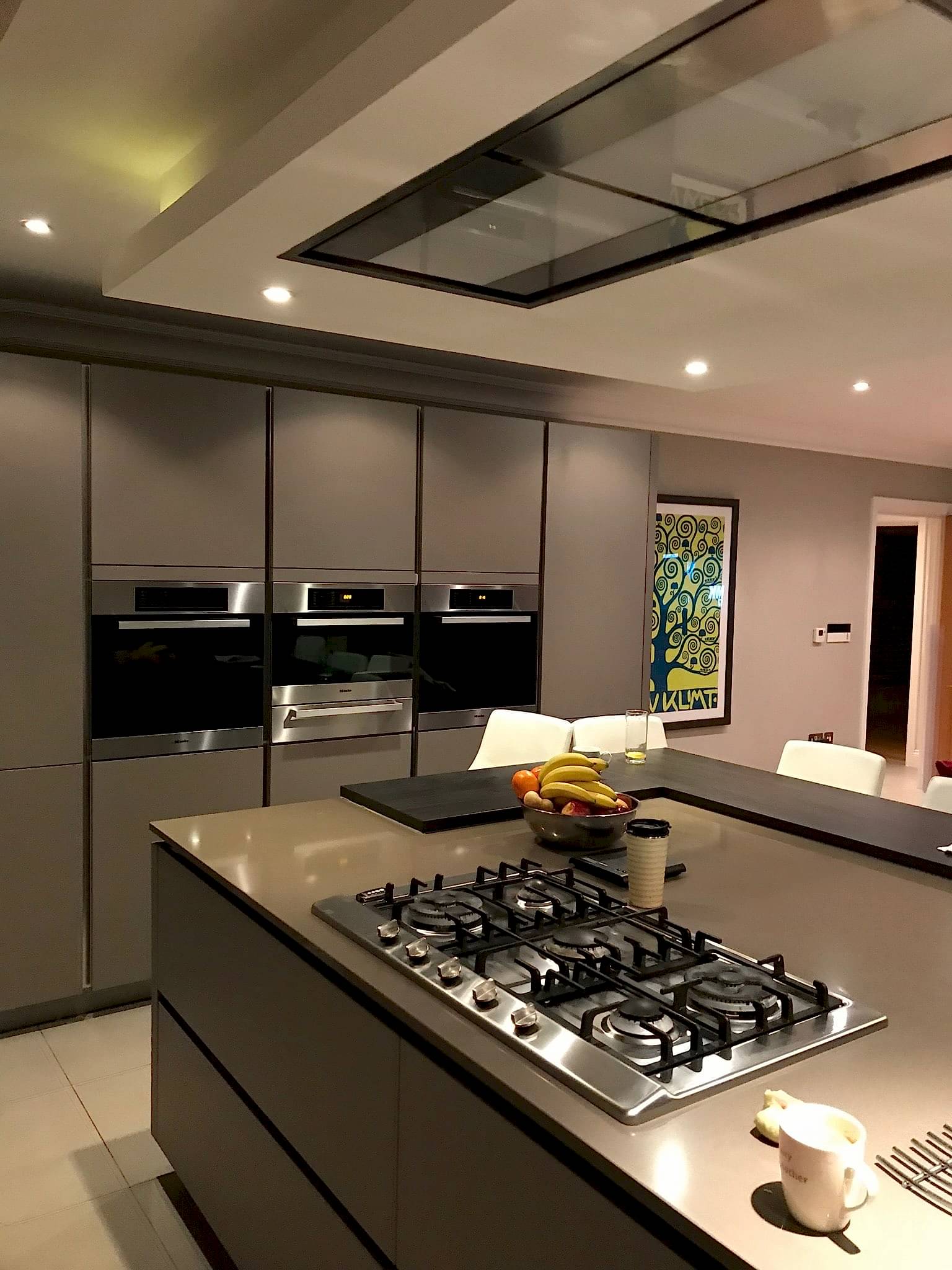 A modern kitchen featuring a large island with a built-in gas cooktop. The island has a fruit bowl, a cup, and a thermos. The background showcases sleek, grey cabinets with built-in ovens and vibrant, framed artwork on the wall. Ceiling lights illuminate the space, creating inviting kitchens for culinary creativity.