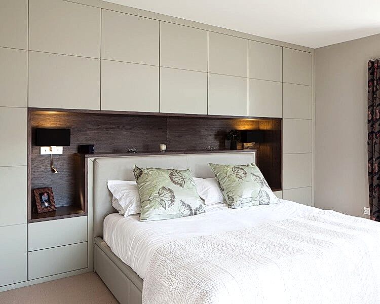 A modern bedroom with a light grey bed and matching fitted furniture. The bed has white bedding and two green pillows with floral patterns. The headboard, part of the fitted cabinetry, creates a recessed shelf with lighting on each side. A framed photo sits on the left shelf, enhancing this elegant fitted bedroom design.
