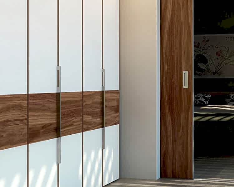 A modern closet door setup featuring white panels with a horizontal wood strip and metal handles brings elegance to your fitted bedroom. Adjacent is a partially opened wooden sliding door, revealing a dimly lit bedroom with a bed and decorative pillows. Sunlight casts shadows on the floor, highlighting the beauty of fitted furniture.