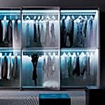 A modern walk-in closet with four sections, each fitted with bespoke wardrobes and illuminated by soft lighting. It contains an array of neatly hung clothes, predominantly in black, white, and gray tones. Two black stools and a stylish lamp are placed in the foreground.
